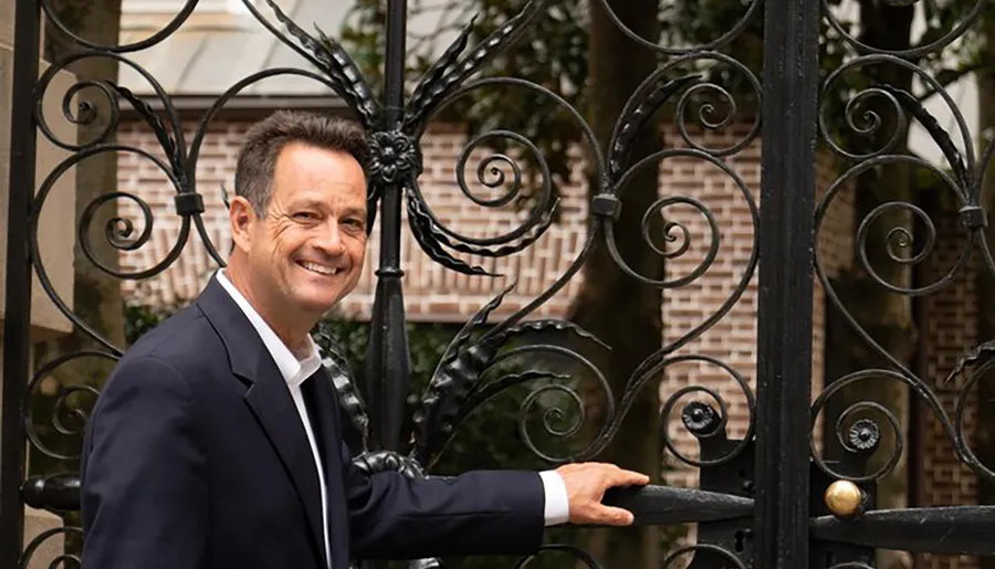 A smiling person in a suit is standing by an ornate wrought-iron gate, with one hand on the gate, posing for the camera.