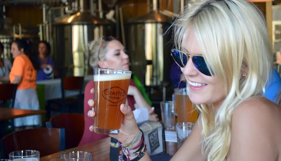 A smiling person in sunglasses is holding up a pint of amber-colored beer inside a brewery with stainless steel tanks visible in the background and other patrons around.