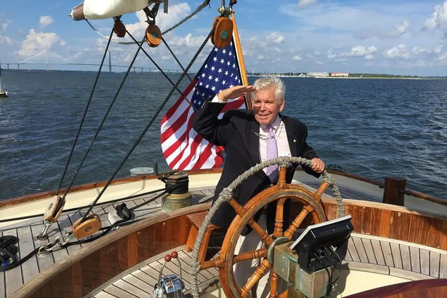 A smiling person is saluting while standing at the helm of a sailboat, with an American flag visible in the background, set against a sunny, cloud-dotted sky over the water.