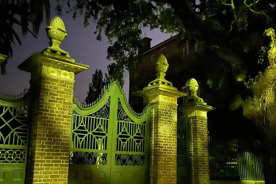 An ornate gate with intricate patterns and statues atop its pillars is cast in a mysterious green light against the night sky.