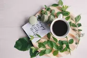 A cup of coffee surrounded by green macarons, fresh leaves, and a note saying 