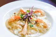 The image shows a gourmet dish consisting of shrimp atop a creamy bed of risotto or a similar grain, garnished with microgreens, served on a white plate.