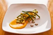 The image shows a gourmet dish consisting of asparagus, broad beans, and sauce artistically plated on a square white plate.