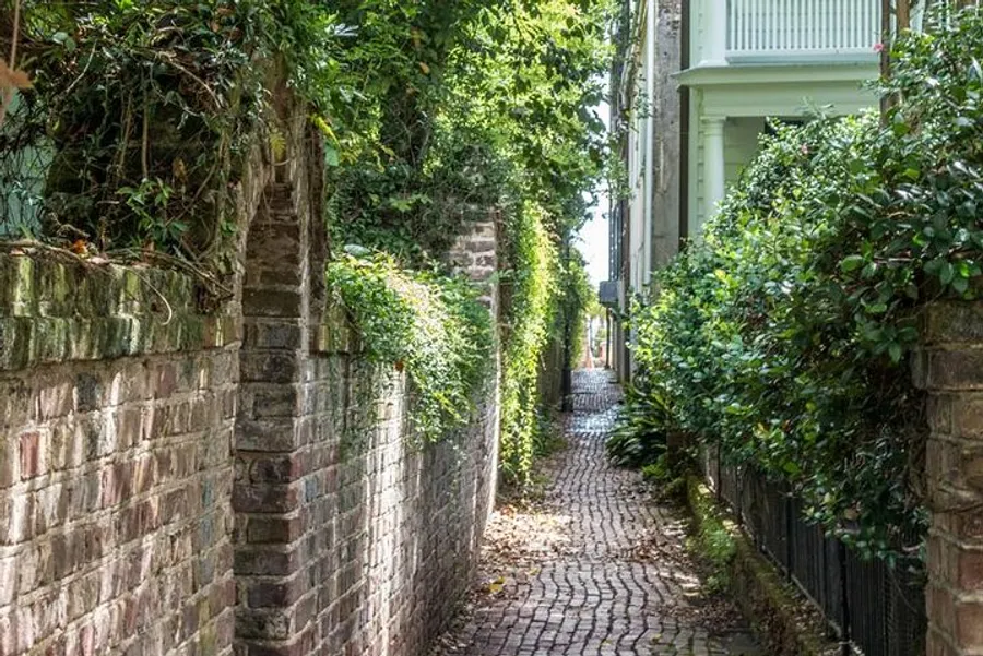 The image shows a narrow cobblestone alley lined with brick walls and lush greenery, creating a quaint and serene path.