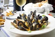 The image displays a plate of cooked mussels garnished with herbs, alongside slices of bread, French fries, a small bowl of sauce, and a glass of white wine.