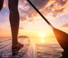 The image captures a close-up of a persons bare feet on a paddleboard with a paddle blade in the water against a beautiful sunset over the ocean