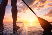 The image captures a close-up of a person's bare feet on a paddleboard, with a paddle blade in the water, against a beautiful sunset over the ocean.