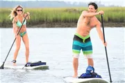 Two people are enjoying paddleboarding in calm waters with smiles on their faces.