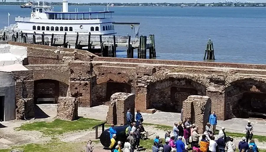 A group of visitors is being guided through the remains of a historic fort with a large boat docked in the background near the water.