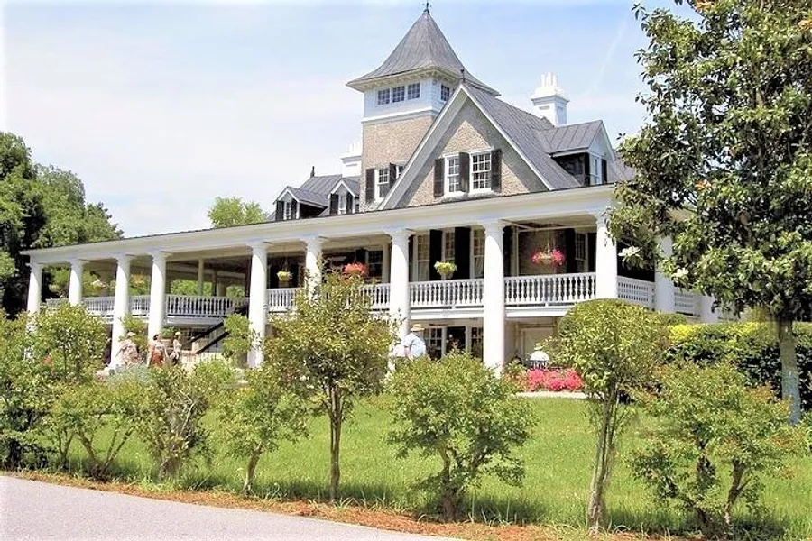 The image features a grand residence with a large front porch and upper balconies, surrounded by well-manicured greenery under a clear sky.
