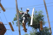 A person wearing a helmet and safety harness is navigating a high ropes course among trees under a clear blue sky.