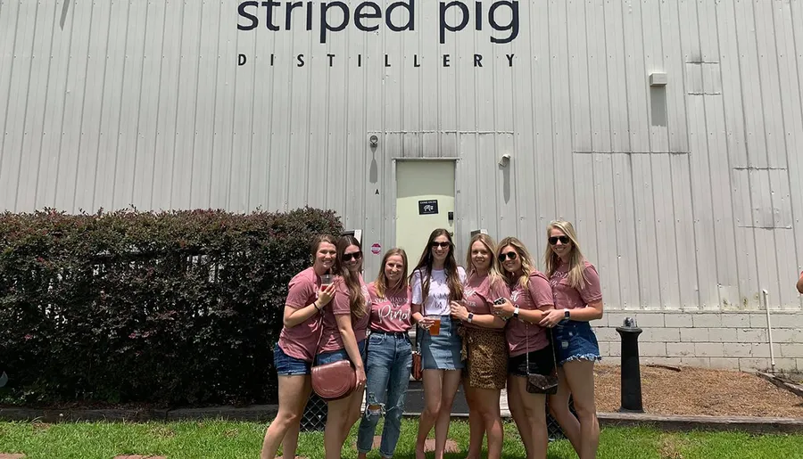 A group of people is posing and smiling in front of the Striped Pig Distillery, with some holding drinks.