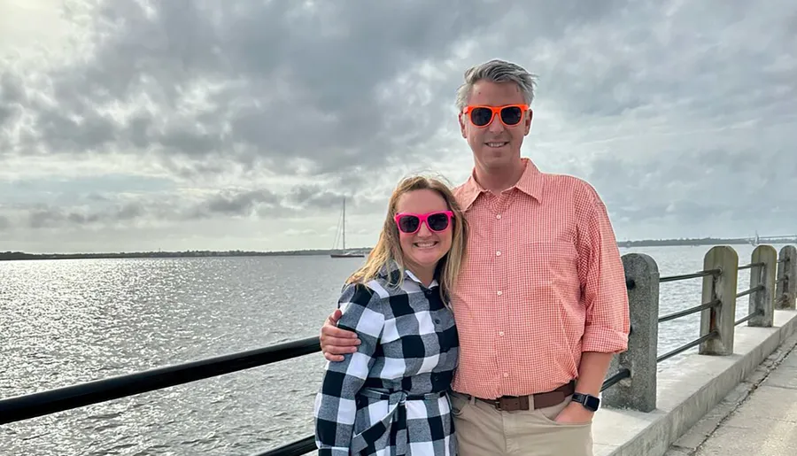 A smiling couple wearing bright sunglasses stands close together with a scenic water view and cloudy sky in the background.
