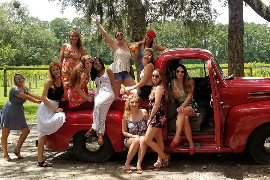 A group of happy women is posing with a vintage red truck in a sunny, outdoor setting.