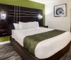 The image shows a modern hotel room with a large bed green accent wall and contemporary furnishings