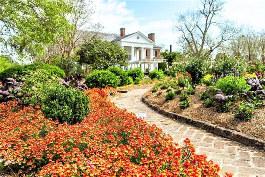 A brick pathway meanders through a vibrant garden leading to a classic white house with large columns.