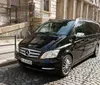 A black Mercedes-Benz V-Class van is parked on the cobblestone street next to a sidewalk and a building with ornate railings