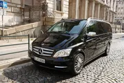 A black Mercedes-Benz V-Class van is parked on the cobblestone street next to a sidewalk and a building with ornate railings.