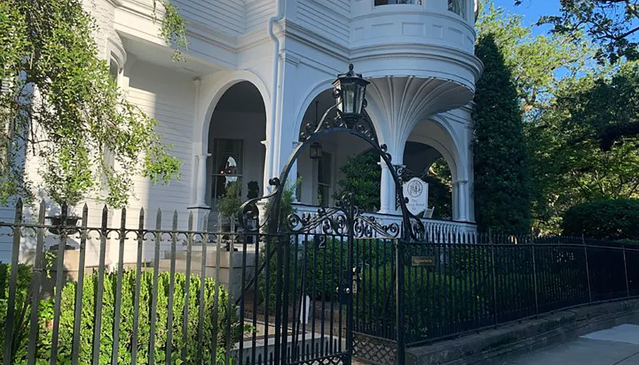 The image shows an elegant white Victorian-style house with a turret, arched entryway, and intricate black iron fence in a lush green setting.