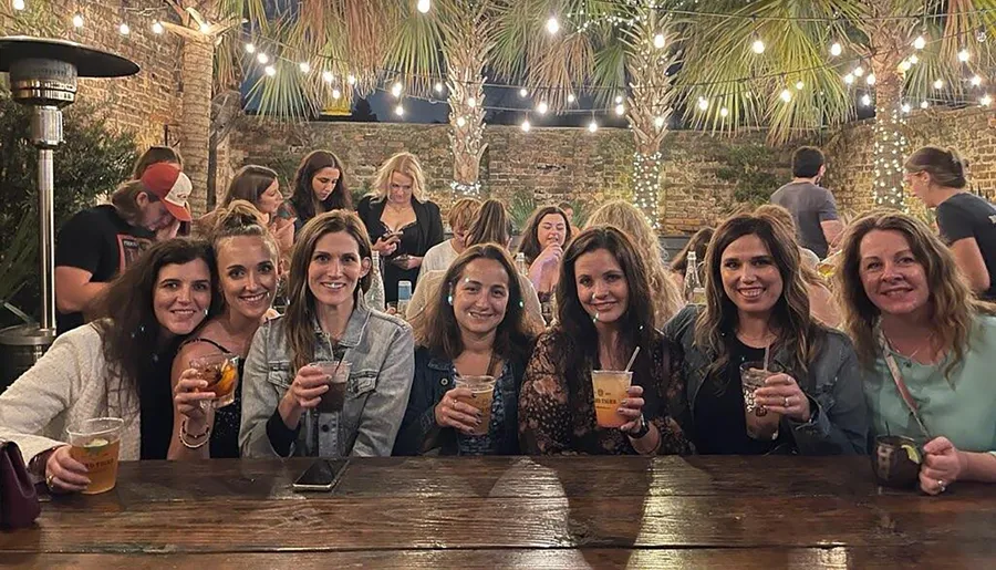 A group of smiling women are gathered at an outdoor table, holding drinks, with a background of fairy lights and a brick wall.