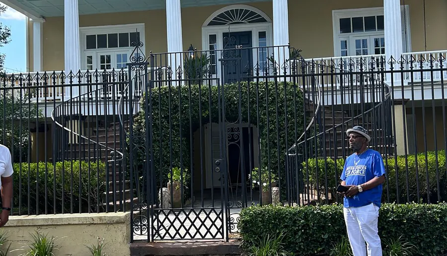A man stands in front of an elegant house with white pillars and a black wrought iron fence on a sunny day.