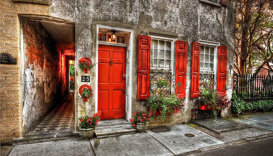 The image shows a charming old building facade with vibrant red doors and shutters, adorned with greenery and flowers, next to a narrow cobblestone alleyway.