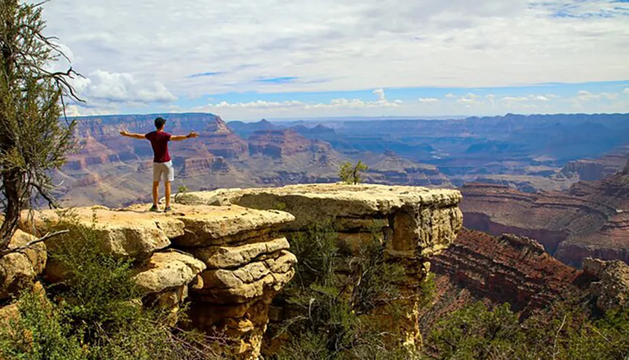 A person stands with outstretched arms on the edge of a cliff overlooking the vast and layered rock formations of the Grand Canyon under a partially cloudy sky.