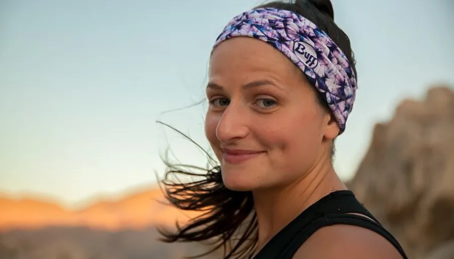 A smiling woman with a headband stands outdoors with a dusk sky and mountain silhouettes in the background.