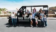 A group of people of various ages posing enthusiastically next to a large, open-air tour vehicle labeled 'BUCK WILD Grand Canyon Hummer Tours.'