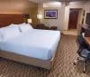 The image shows a neatly arranged hotel room with a king-sized bed a work desk with a chair and a flat-screen TV