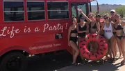 A group of people in swimwear is posing cheerfully beside a red bus with the slogan 
