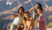 Four friends are taking a group selfie with a smartphone on a selfie stick, with a scenic canyon backdrop illuminated by sunlight.