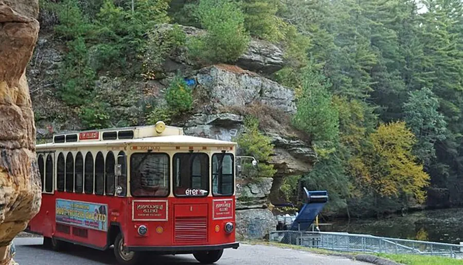 A red trolley bus with advertisements on its side is driving by a rocky outcrop surrounded by lush greenery and a few trees with yellow autumn leaves.