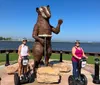 Two people on Segways are posing next to a large metal statue of a bear by a lakeside