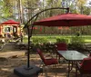 This image depicts a peaceful outdoor patio area with red cushioned chairs a table under a red umbrella and a grill all set on flagstone paving with a lush green background of a well-kept garden and trees