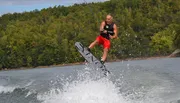 A person is captured mid-air while wakeboarding on a lake with a backdrop of trees.