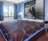 The image shows a neatly arranged hotel room with a large bed featuring a blue and brown geometric bedspread a matching curtain a big black and white nature photograph on the wall and basic furniture like a desk chair and nightstands