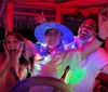 Three people are joyfully posing with hands up around a wooden ships steering wheel under pink lighting with one wearing a blue hat and lei suggesting a festive or party atmosphere