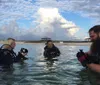 Three scuba divers are preparing for a dive in shallow water near the shore under a sky with large cumulus clouds