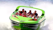 A group of people are experiencing a high-speed thrill ride on a boat designed to look like a menacing green creature with sharp teeth, spraying water behind as it races forward.