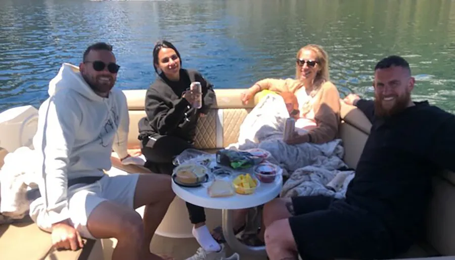 Four people are casually enjoying a sunny day on a boat, with snacks and drinks laid out on a table between them.