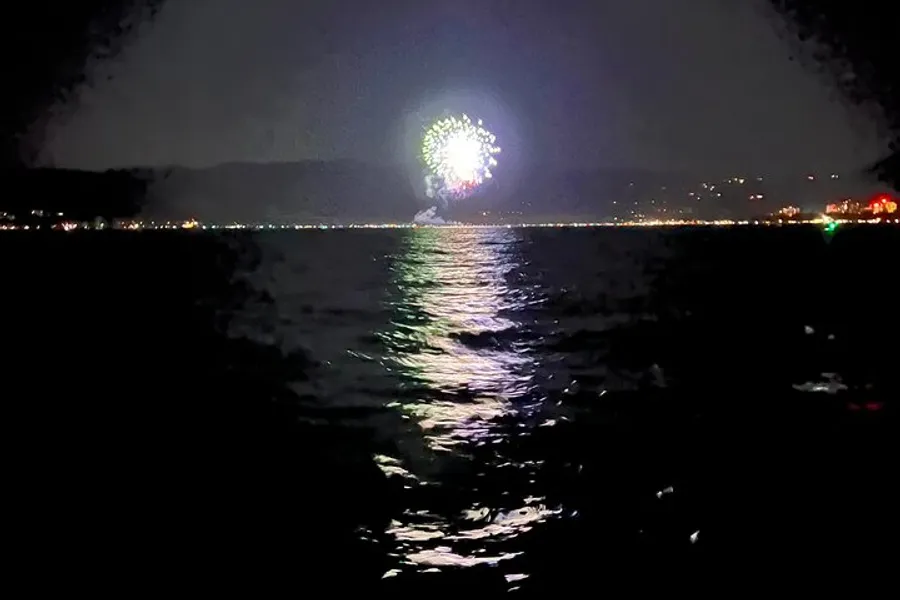 A firework bursts in the night sky over a body of water, with its reflection shimmering on the surface.
