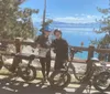 Two individuals in cycling attire are posing with their bicycles in front of a scenic lake view