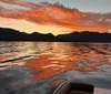 A vibrant sunset paints the sky with shades of orange and red above a serene mountain-lined lake as seen from the edge of a boat