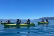 A group of people are kayaking on calm blue water with a backdrop of majestic snow-capped mountains under a clear blue sky.