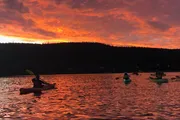 A group of kayakers paddle on a lake under a vivid orange sunset sky.