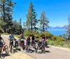 A group of four cyclists one being a young child are riding on a paved trail with a scenic forest landscape and a lake in the background under a clear blue sky