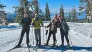 Four people are smiling for the camera while standing on a snowy slope with cross-country skis, against a backdrop of snow-covered trees and a blue lake in the distance.