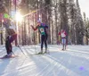 Three people are cross-country skiing among snowy trees with the sun low on the horizon creating long shadows and a flare effect in the photo