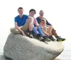 The image shows four individuals of varying ages sitting closely together atop a large rock with a seemingly calm body of water and a hazy sky in the background